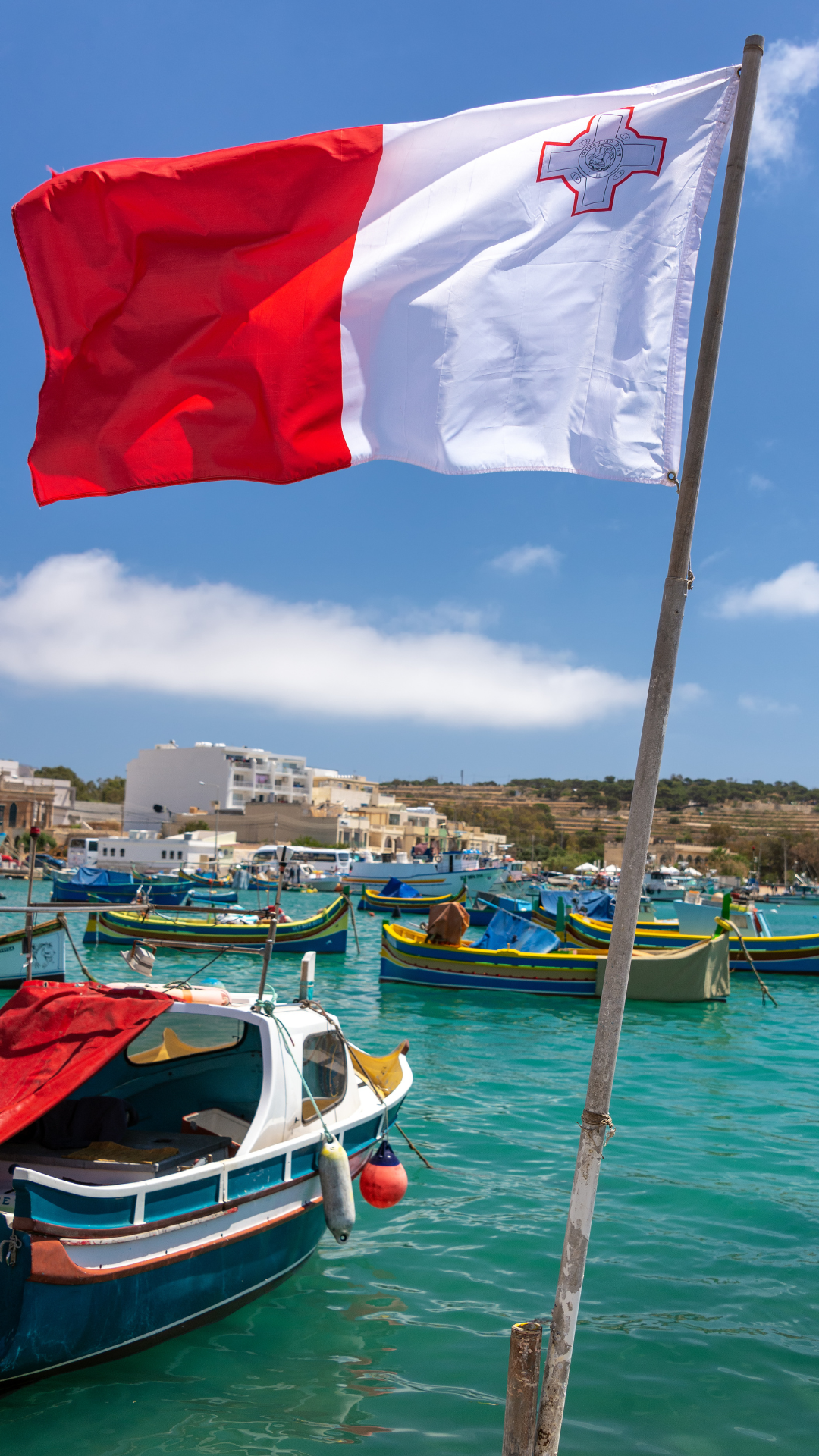 Hier siest du wie die Malta Flage mit ihren typischen Farben, rot und weiß im Wind weht. Im Hintergrund siehst du einen Hafen mit einem kleinen Bot und türkisem Wasser.