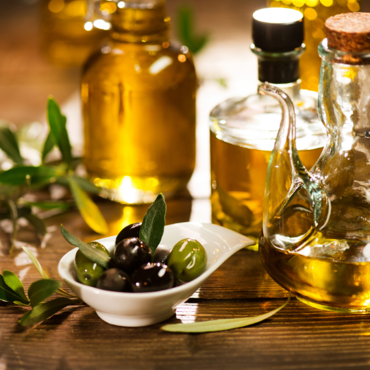 Olivenöl gehört in jedes Italien Essen. Schau dir das hochwertige italienische Olivenöl auf dem Bild an. Es ist in Flaschen Gefüllt und steht auf einem Tisch neben Oliven in einer kleinen weißen Schale.